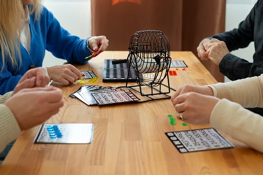 Cztery osoby grają w bingo przy drewnianym stole. Na stole znajduje się szafka do bingo oraz kolorowe żetony, a atmosfera jest wesoła i spokojna.