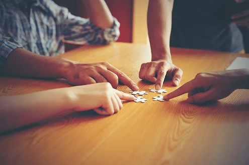 Ręce czterech różnych osób wskazują na rozsypane kawałki układanki na drewnianym stole, symbolizując pracę zespołową i współpracę. Miękkie oświetlenie tworzy ciepłą atmosferę.